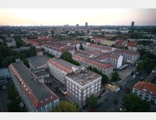 Luftaufnahme eines städtischen Wohngebiets mit mehreren Gebäuden im Stil der Nachkriegsarchitektur, gekennzeichnet durch rote Ziegeldächer und helle Fassaden. Im Vordergrund befindet sich ein Flachbau mit bepflanzter Dachterrasse, auf der sich eine Gruppe von Menschen versammelt hat; umliegende Straßen und Bäume rahmen die Szene ein.