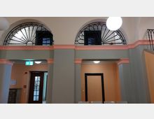 Innenansicht eines Gebäudes mit bogenförmigen Fenstern, die mit dekorativen schwarzen Gitterelementen versehen sind. Der Raum ist in hellen Pastelltönen gestaltet, mit orangefarbenen Wänden, grauen Säulen und rosa Zierleisten, ergänzt durch helle Beleuchtung und ein Notausgangsschild.