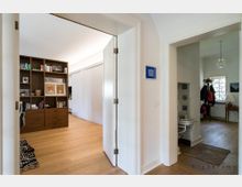 Heller Innenraum eines Hauses mit Blick auf zwei Räume: Links ein Arbeitszimmer mit offenem Regal und Holzboden, rechts ein Eingangsbereich mit Garderobe, Spiegel und Waschbecken, beleuchtet durch ein Fenster mit Gittermuster.