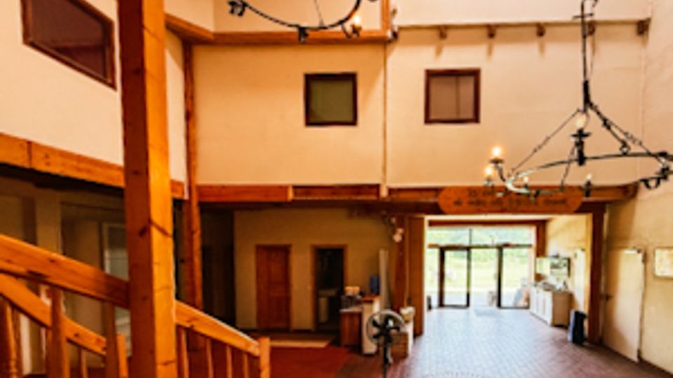 Innenansicht eines rustikalen Saals mit sichtbaren Holzbalken, Treppe mit Holzgeländer und Ziegelfliesenboden. An der Decke hängen mehrere große Kronleuchter, und am Ende des Raums befinden sich Türen und große Fensterfronten.