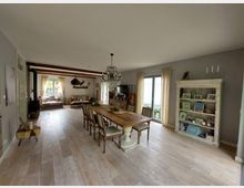 Wohnzimmer mit Essbereich in einem rustikalen Stil. Es gibt einen langen Holztisch mit Stühlen, eine helle Sitzecke mit großen Fenstern, einen Kronleuchter und ein weißes Regal mit dekorativen Gegenständen.