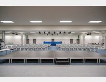 Konferenzraum mit einer leeren Bühne im Vordergrund, Stuhlreihen und einem blauen Rednerpult im Hintergrund. Die Decke ist mit Deckenleuchten und Hängekonstruktionen für Lichttechnik ausgestattet, die Wände sind neutral gestaltet.