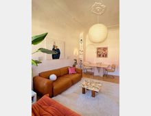 Wohnzimmer mit heller, dekorativer Stuckdecke und modernem Interieur. Zu sehen sind ein braunes Ledersofa, ein Couchtisch mit Marmorplatte, Pflanzen, abstrakte Wandbilder sowie ein Essbereich mit Holztisch und Stühlen im Hintergrund.