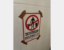 Ein Plakat mit der Aufschrift „Toilettenbenutzung nur für Menschen“ und der Werbung für den Film „District 9“ ist an einer weißen Wand befestigt. Es zeigt eine durchgestrichene Silhouette eines außerirdischen Wesens und ist mit braunem Klebeband fixiert.