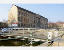 Ehemaliges, mehrstöckiges, historisches Gebäude mit verblasster Fassade und beschädigten Fenstern, im Vordergrund eine kleine Brücke über einen Kanal sowie parkende Autos links. Die Umgebung ist urban und von kahlen Bäumen geprägt.