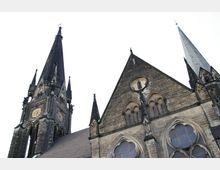 Kirchenfassade mit gotischem Design, bestehend aus hohen, spitzen Türmen, großen Spitzbogenfenstern und dekorativen Details aus dunklem Stein; links eine Uhr auf einem Turm, im Hintergrund ein heller, wolkenloser Himmel.