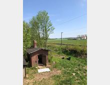 Backsteinbau mit offen stehender Tür in einer ländlichen Umgebung, umgeben von Bäumen, Wiesen und einem Bahngleis im Hintergrund. In der Ferne sind ein Wohnhaus und eine weite grüne Landschaft zu sehen.