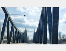 Moderne Stahlbrücke mit geschwungenen, blauen Trägern, die sich in regelmäßigen Abständen wiederholen. Im Hintergrund sind Bäume, ein Obelisk und ein bewölkter Himmel zu sehen.