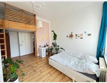 Helles Schlafzimmer mit Holzboden und einer hohen Decke, ausgestattet mit einem weißen Bett, einem kleinen Schreibtisch und mehreren Zimmerpflanzen. Ein Hochbett aus Holz mit Leiter befindet sich über einer Tür im Hintergrund, und die weiße Wand ist dezent mit Dekorationen wie Fotos und Zeichnungen geschmückt.