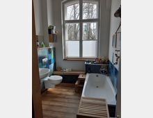 Badezimmer mit einem großen Fenster, das Blick auf kahle Bäume bietet. Der Raum hat Holzboden, eine Badewanne, ein Waschbecken und eine Toilette, sowie blaue und grüne Wandfliesen um die Badewanne herum.