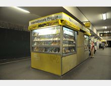 Ein Zeitschriften- und Tabakwarenkiosk in einer U-Bahn-Station mit gelbem Schild und der Aufschrift „Internationale Presse“. Der Kiosk ist von Zeitungen und Magazinen in beleuchteten Regalen umgeben, die Bahnsteigumgebung ist mit Sitzmöglichkeiten und einer Uhr im Hintergrund ausgestattet.