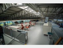 Industriehalle mit Zwischengeschoss, das als Aufenthaltsbereich genutzt wird. Mehrere Tische mit karierten Tischdecken und roten Stühlen, umgeben von Metallgeländern, sind unter einem Dach aus Stahlträgern angeordnet; im Hintergrund sichtbar sind Arbeitsbereiche und Lagerregale.