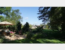 Großer Garten mit grünem Rasen, üppiger Bepflanzung und einem Sitzbereich mit Sonnenschirm und Korbmöbeln. Im Hintergrund befindet sich ein helles Gebäude, umgeben von Bäumen und Sträuchern.