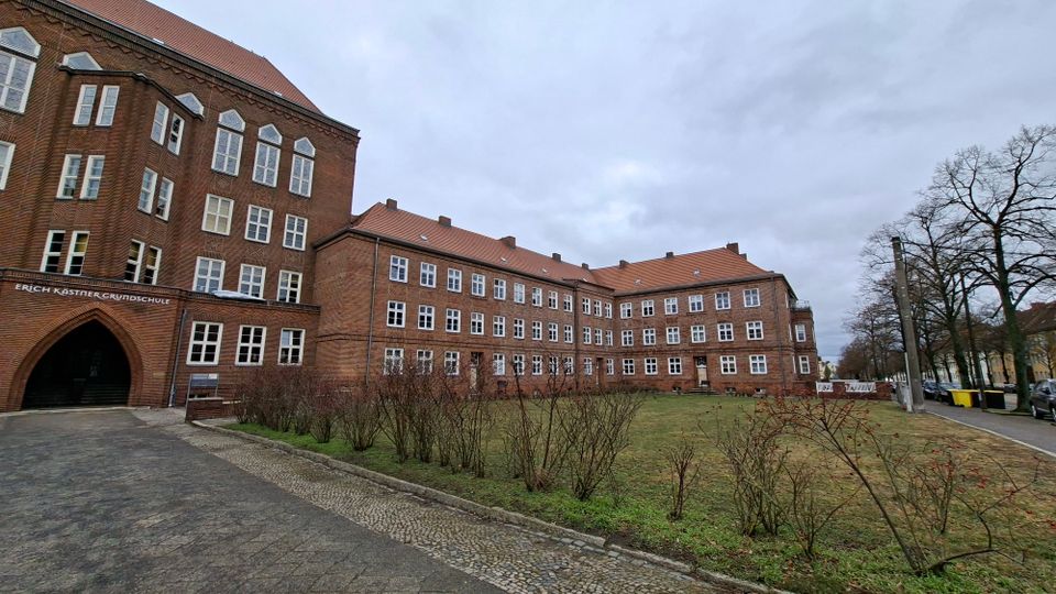 Großes, mehrstöckiges Backsteinschulgebäude mit roten Ziegeldächern und vielen symmetrisch angeordneten Fenstern, links mit einem spitzbogigen Eingang. Davor liegen ein gepflasterter Weg und eine Rasenfläche mit niedrigen Sträuchern; am Rand stehen kahle Bäume und eine Straße.
