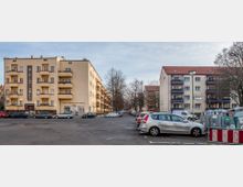 Wohngebiet mit mehrstöckigen, beige- und cremefarbenen Wohnhäusern, einige mit Balkonen. Der Vordergrund zeigt parkende Autos auf einem Parkplatz, rechts stehen Müllcontainer, und Bäume säumen den Straßenrand im Hintergrund.