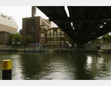 Backstein-Gebäude im Stil des Industriezeitalters an einem Flussufer, mit Schornstein und detaillierten Verzierungen. Im Vordergrund ist das Wasser zu sehen, während ein Brückengerüst rechts den Blick zum Teil überspannt.