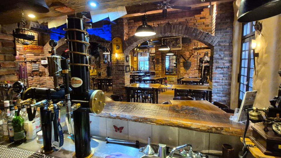 Innenansicht einer rustikalen Bar bzw. Kneipe mit freiliegenden Backsteinwänden, Rundbögen und warmem Deckenlicht. Im Vordergrund steht ein Holztresen mit Zapfanlage und Flaschen, dahinter ein Sitzbereich mit Holztischen und -stühlen sowie dekorativen Vintage-Objekten.