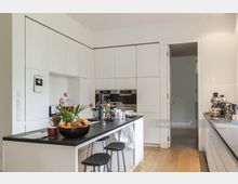 Moderne Küche mit weißen, deckenhohen Einbauschränken, einer schwarzen Arbeitsplatte und Holzboden. Auf der Kücheninsel stehen eine Obstschale, frische Tulpen und Haushaltsgeräte; im Hintergrund führt eine Tür zu einem angrenzenden Raum.