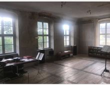 Ein heller Raum mit großen, weiß gerahmten Sprossenfenstern, einem Holzboden und sichtbaren Putzwänden. Im Raum befinden sich ein Tisch mit Stühlen, Musiknoten und ein Notenständer sowie Regale an den Wänden.