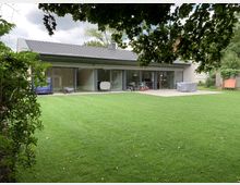 Ein modernes Wohnhaus mit Flachdach, großen bodentiefen Glasschiebetüren und einem weitläufigen Gartenrasen im Vordergrund. Links und rechts befinden sich Bäume und Sträucher, die den Garten umrahmen.