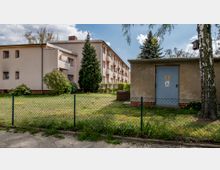 Ein zweistöckiges Wohngebäude mit beiger Fassade und Balkonen, daneben ein kleines, fensterloses Technikgebäude mit metallener Tür, umgeben von einem Zaun und einer Rasenfläche mit vereinzelten Bäumen und Sträuchern im Vordergrund.