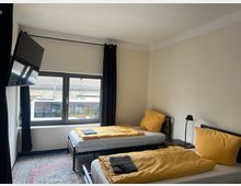 Schlichtes Hotelzimmer mit zwei Einzelbetten, gelber Bettwäsche und einem Nachttisch mit kleiner Lampe. Ein großes Fenster mit schwarzen Vorhängen und ein an der Wand montierter Flachbildfernseher sind sichtbar.