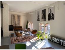 Heller Raum im Loft-Stil mit weißen Wänden, großen runden Fenstern und industriellen Elementen. Der Raum ist mit Stühlen, einem Sofa, Teppichen und Pflanzen eingerichtet; an der Wand hängen großformatige Schwarz-Weiß-Fotografien.