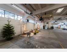 Industriehalle mit moderner Einrichtung: Große, helle Halle mit weißen Wänden und Dachbalken, teilweise möbliert mit Sitzgruppen aus Stühlen und bunten Hockern. Ein geschmückter Weihnachtsbaum steht links, weitere Dekorationen und Tische befinden sich im Hintergrund.