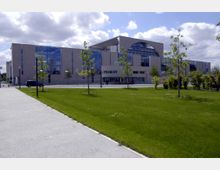 Modernes Regierungsgebäude mit großen Glasfassaden und einem markanten halbrunden Fenster im oberen Bereich. Im Vordergrund ein gepflegter Rasen mit jungen Bäumen sowie ein Gehweg, darüber ein leicht bewölkter Himmel.