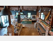 Innenraum eines rustikalen Hofladens mit Backsteinwänden, sichtbaren Holzträgern und großen Decklampen. Zu sehen sind Regale mit Lebensmitteln, ein Tresen mit der Aufschrift "Regional Markthalle" und eine Selbstbedienungstheke mit Schüttbehältern für lose Waren.