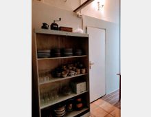 Ein einfaches Regal in einer Küche oder Abstellkammer mit Geschirr: Teller, Tassen, Gläser und Behälter verteilt auf verschiedenen Regalböden. Oben befinden sich dekorative Gegenstände, wie eine Kaffeemühle, eine Vase und eine Glaskuppel; rechts ist eine geschlossene weiße Tür zu sehen.