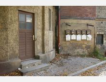 Eine alte Gebäudeecke mit bröckelndem Putz, einer braunen Holztür und mehreren Briefkästen an der Wand; der Boden ist mit Kopfsteinpflaster und Laub bedeckt.