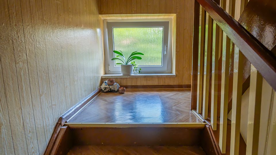 Treppenhaus-Innenraum mit holzvertäfelten Wänden und glänzendem Parkettboden, rechts eine Treppe mit dunklem Handlauf und hellen Geländern. Am Ende des Podests befindet sich ein Fenster mit Milchglas, auf der Fensterbank stehen Topfpflanzen und kleine Dekorationsgegenstände.