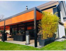 Moderne Hausfassade mit überdachter Terrasse, schwarzem Metallrahmen und orangefarbener Markise vor großen Glasschiebetüren. Daneben stehen ein hoher Bambus im Pflanzkübel, Terrassenmöbel und ein Rasen mit Trittsteinen.