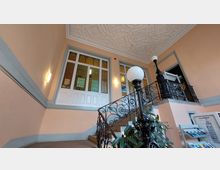 Treppenhaus eines historischen Gebäudes mit verzierten Metallgeländern, großen Buntglasfenstern und einer dekorativen Stuckdecke. Im Vordergrund befindet sich eine antike Lampenhalterung und eine Pflanze, im Hintergrund ein Prospektständer.