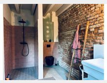Innenansicht eines schmalen Badezimmers mit freiliegender Ziegelsteinwand, terrakottafarbenen Bodenfliesen und einer Dusche mit schwarzer Armatur. Eine Holzleiter mit gestreiftem Handtuch lehnt an der Wand; daneben stehen ein schwarzes Waschbecken und eine Badewanne.