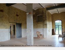 Erdgeschoss eines historischen Gebäudes mit Backsteinwänden, hohen Decken und einem Betonpfeiler in der Mitte. Im Raum stehen vereinzelte Stühle, und durch die Glastür im Hintergrund fällt Blick auf eine grüne Außenfläche.
