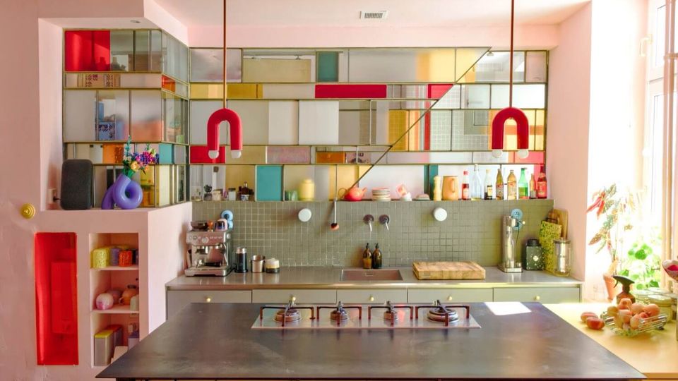 Bright modern kitchen with a large central metal island and built-in stovetop, backed by gray tile counters and shelves lined with bottles and small appliances. A wall of colorful stained-glass-style panels and two red U-shaped pendant lights frame the space, with sunlight coming in from tall windows on the right.