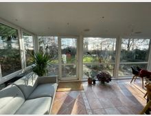 Wohnbereich in einem Wintergarten mit großen Glasfenstern, die einen Blick auf einen Garten mit Bäumen und Pflanzen bieten. Innen befinden sich ein graues Sofa, eine Topfpflanze, einige kleinere Pflanzen auf dem Boden und Fliesenboden mit dekorativen Mustern.