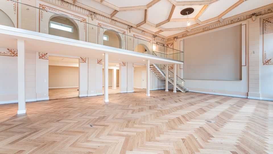 Heller, leerer Saal mit großem Fischgrät-Parkett und einer umlaufenden Empore mit Glasgeländer und Treppe. Die hohe, reich verzierte Kassettendecke sowie Rundbögen und Wandstuck prägen den historischen Innenraum.
