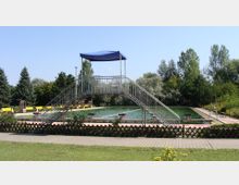 Freibad mit einem rechteckigen Schwimmbecken, umgeben von Bäumen und gelben Bänken. Im Vordergrund ist eine erhöhte Plattform mit blauer Überdachung und Treppen auf beiden Seiten sichtbar.