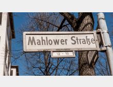 Straßenschild mit der Aufschrift „Mahlower Straße“ und den Hausnummern „12–16“, befestigt an einem Metallpfosten. Im Hintergrund sind ein Baum mit kahlen Ästen und ein Teil eines Gebäudes zu sehen, unter einem klaren blauen Himmel.