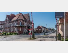 Straßenansicht in Neuruppin mit einem historischen Backsteingebäude mit rotem Satteldach im Vordergrund, einer beschrankten Bahnübergangsanlage und dem Straßenschild "Neuruppin Rheinsberger Tor". Im Hintergrund sind weitere Gebäude und eine gerade verlaufende Straße zu sehen.