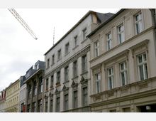 Straßenszene mit historischen Wohnhäusern, die verputzt oder aus Ziegelstein gebaut sind und dekorative Fassadenelemente wie Gesimse und Fensterrahmungen aufweisen; links ist ein Baukran sichtbar.