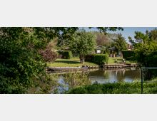 Grüne Uferlandschaft mit einem kleinen Fluss, an dem ein Holzsteg und ein Boot zu sehen sind. Im Hintergrund befinden sich Rasenflächen, Wohnhäuser mit Gärten sowie Bäume und Sträucher.