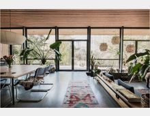 Wohnraum mit modernem, offenem Design, großen Glasfronten und Blick auf einen Garten. Innenraum mit Holzelementen an der Decke, Pflanzen, einem langen Esstisch, Sesseln und buntem Teppich auf hellem Boden.