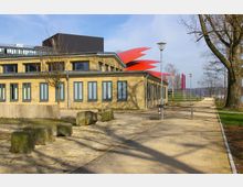 Backsteingebäude mit großen Fenstern, umgeben von einem gepflasterten Weg und Grünflächen; auffällige rote, geschwungene Dachkonstruktion im Hintergrund. Im Vordergrund liegen vereinzelt größere Steinblöcke, und links steht ein einzelner Baum.