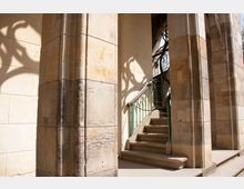 Steinernes Treppenhaus an einem historischen Gebäude mit hellen Sandsteinwänden und gotischen Elementen. Eine grüne Metallgeländerbiegung und verspielte Schatten auf der Wand betonen die dekorative Gestaltung.