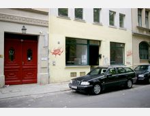 Straßenszene mit einer gelb-beigen Hausfassade, einem roten Doppeltor links und Schaufenstern rechts, teilweise mit Graffiti bedeckt. Auf der Straße parken vorne in der Bildmitte ein schwarzer Kombiwagen und ein weiteres Auto.