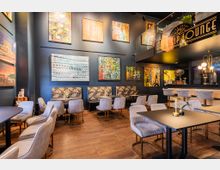 Moderne Lounge mit einem künstlerischen Ambiente, ausgestattet mit grauen Polsterstühlen, floralen Sitzbänken und dunklen Holztischen. Die Wände sind mit abstrakten Gemälden geschmückt, beleuchtet von warmem Licht, und eine Galerieebene sowie eine Bar sind im Hintergrund sichtbar.